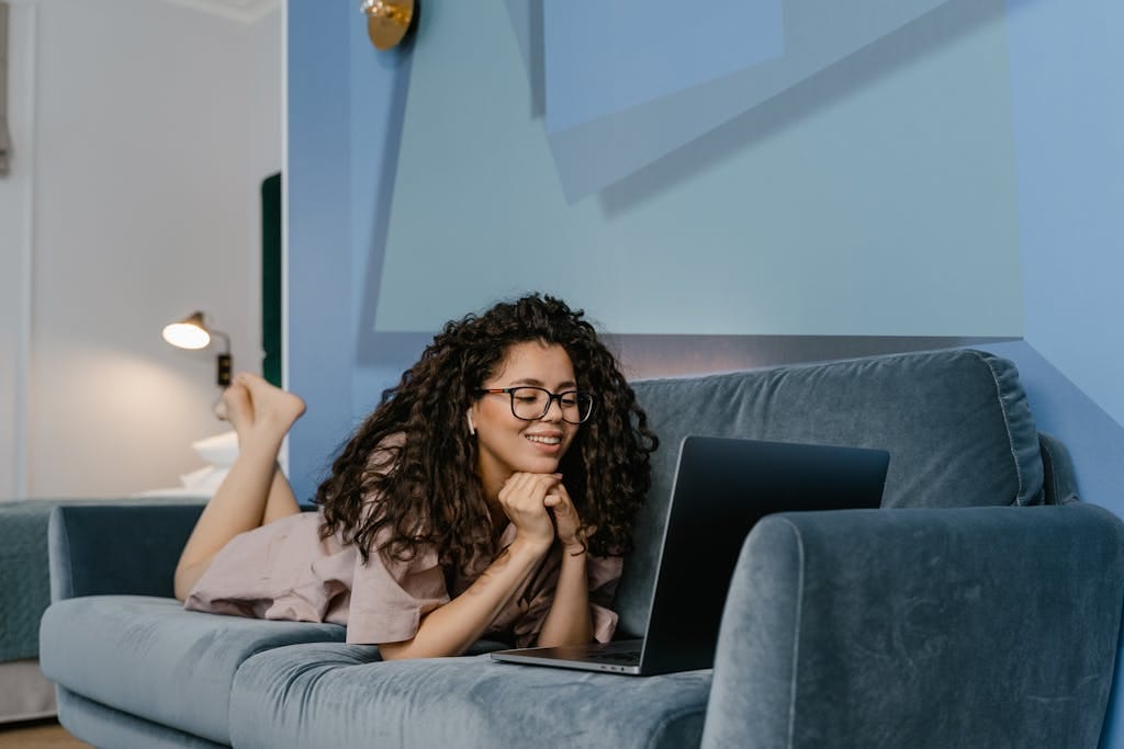 Happy woman with curly hair video calling on laptop, lying on sofa in cozy living room.