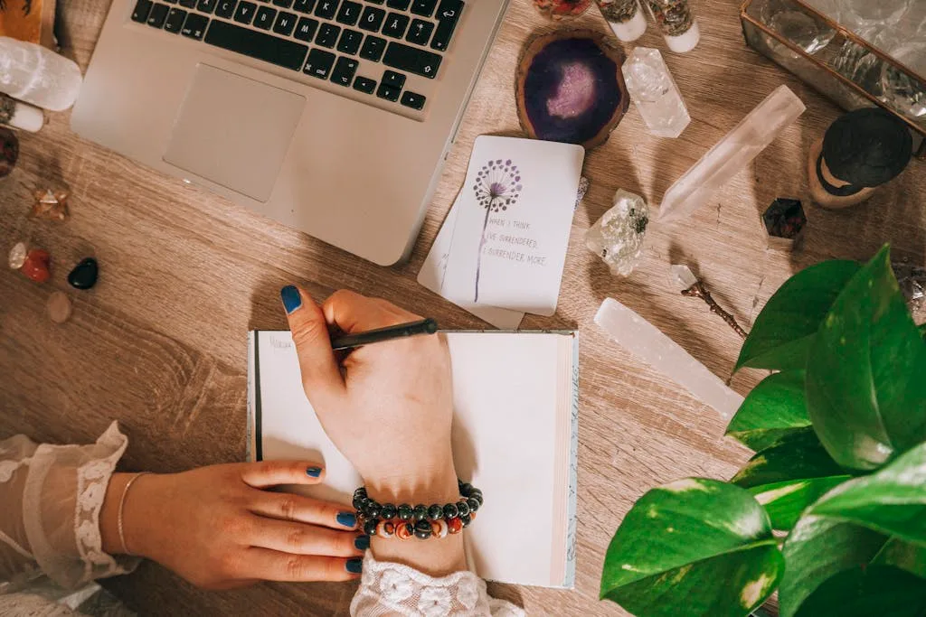 Woman's hand writing in a journal surrounded by crystals, plants, and laptop on a wooden desk.